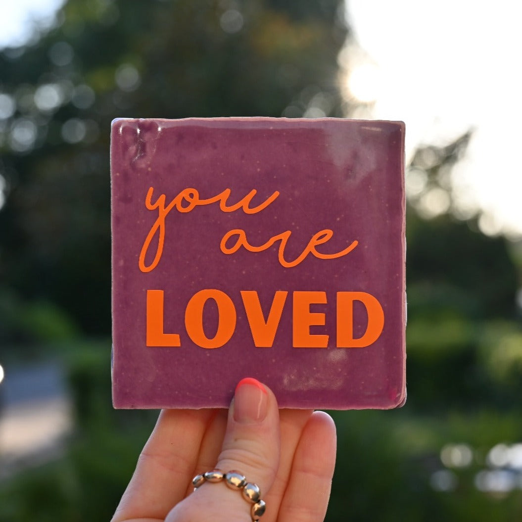 Mooie standaard quote tegel met tekst "You are loved" in een hand. Tegelkleur paars met oranje tekst opdruk. Schitterend in het licht.